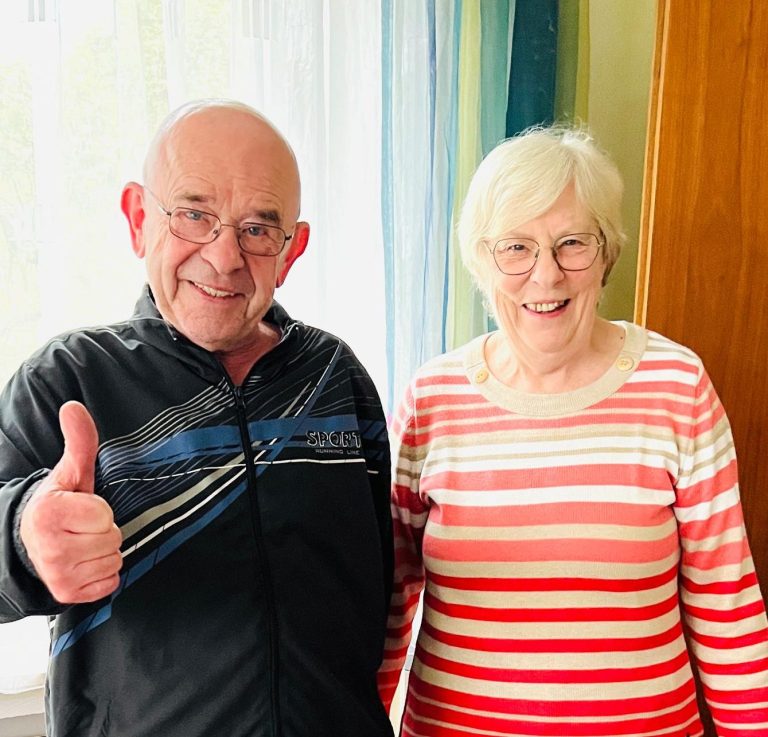 Ein älterer Mann gibt ein Daumen-hoch, neben einer Frau mit Brille in gestreiftem Shirt.