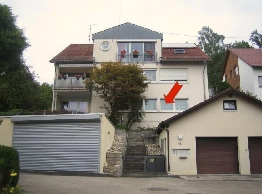 Zweifamilienhaus mit grau-blauen Fensterläden, einem Anbau und Garage. Rote Pfeilmarkierung auf Fenster.