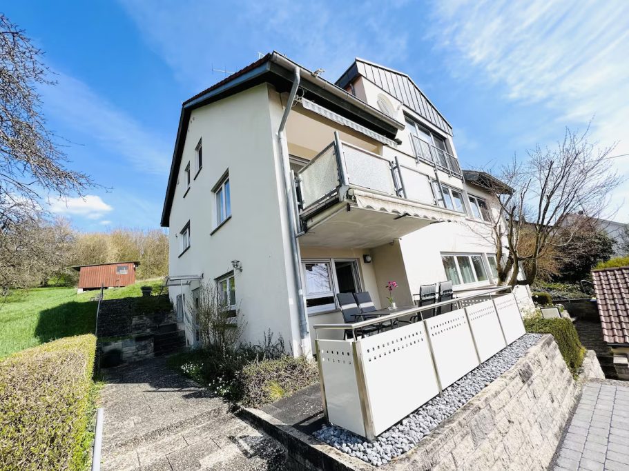 Modernes Gebäude mit Balkon, umgeben von Grünflächen und blauem Himmel.